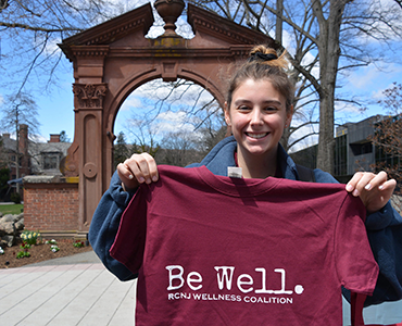 Student standing at the arch holding a Be Well t shirt